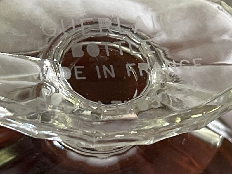 May include: Close-up of a clear glass bottle base with the text "GUERLAIN BOTTLE MADE IN FRANCE". The bottle has a geometric design and a brown liquid inside.