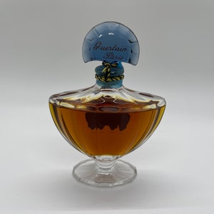 May include: A vintage perfume bottle with a clear glass base and a rounded, amber-colored body. The bottle has a blue, fan-shaped stopper with "Guerlain Paris" printed on it. The stopper is secured with a gold and blue cord.
