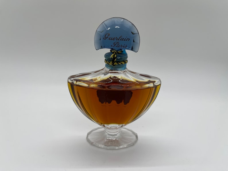 May include: A vintage perfume bottle with a clear glass base and a rounded, amber-colored body. The bottle has a blue, fan-shaped stopper with "Guerlain Paris" printed on it. The stopper is secured with a gold and blue cord.