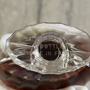 May include: Close-up of a clear glass perfume bottle with a faceted, oval-shaped stopper. The bottle has a brown liquid inside. The text "BOTTLE MADE IN FRANCE" is etched on the stopper.