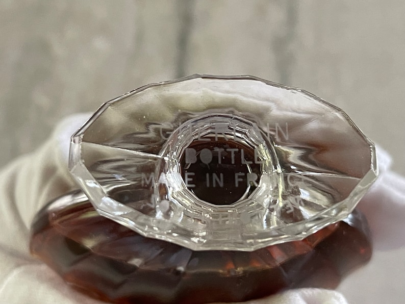 May include: Close-up of a clear glass perfume bottle with a faceted, oval-shaped stopper. The bottle has a brown liquid inside. The text "BOTTLE MADE IN FRANCE" is etched on the stopper.