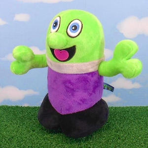 May include: A plush toy character with a green head, arms, and nose, blue eyes, and a pink heart-shaped mouth. It wears a purple shirt, a gray band around its neck, and black pants. The toy is on a green surface with a blue sky background.