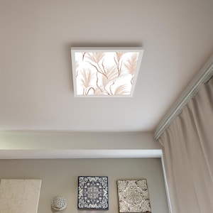 May include: A square, white ceiling light fitting with a decorative panel featuring a pattern of beige and brown feathery plant designs on a white background. The light is mounted on a white ceiling.