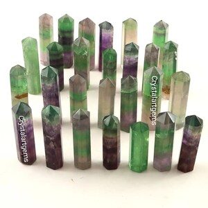 May include: A collection of fluorite crystal points in various shades of green, purple, and clear. The crystals are cut into a hexagonal shape and are arranged on a white surface. The text "Crystalartgems" is visible on some of the crystals.