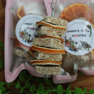 May include: A treat skewer featuring alternating layers of rectangular beige oat blocks and dried orange slices. The skewer is suspended in front of two clear plastic pouches, each labeled with a circular sticker reading AMBER'S FLOCK GOODIES.