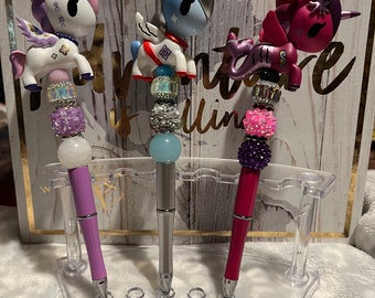 May include: Three unicorn-themed pens with colorful beaded accents. The pens are displayed in a clear plastic pen holder. The unicorn toppers are pink, blue, and purple.