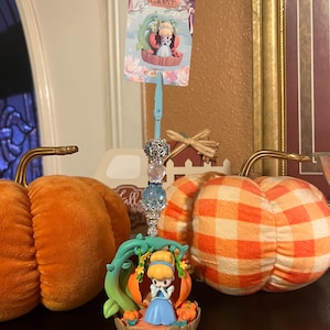 Puede incluir: Una figura de Cenicienta con un vestido azul y un carruaje de calabaza. La figura forma parte de una exhibición decorativa, con calabazas naranjas y una tarjeta con texto japonés. La exhibición tiene un tallo azul con cuentas decorativas.