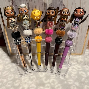 以下が含まれることがあります： 異なる漫画のキャラクターのトッパーが付いた6本のカラフルなペン。ペンは透明なプラスチックスタンドに表示されています。トッパーには、プリンセス、ひげを生やした男性、黄色のキャラクター、茶色のキャラクター、茶色の犬、茶色の髪の女の子が含まれています。