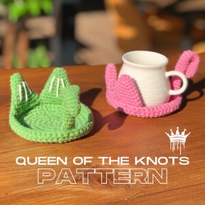 Könnte beinhalten: Zwei gehäkelte Untersetzer in Form von Kronen. Einer ist grün mit weißen Akzenten und der andere ist rosa. Die Untersetzer befinden sich auf einer Holzoberfläche. Der Text "Queen of the Knots Pattern" befindet sich unter den Untersetzern.