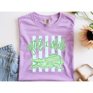 May include: A lilac-colored t-shirt with the text "after a while" in a cursive font, along with a green alligator graphic. The design includes vertical light blue stripes. The shirt is folded and placed on a white surface.