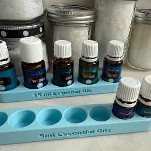May include: A display of essential oil bottles in a light blue holder. The bottles are brown with white caps and blue labels. The holder has the text "15 ml Essential Oils" and "5ml Essential Oils". Jars of white powder are in the background.