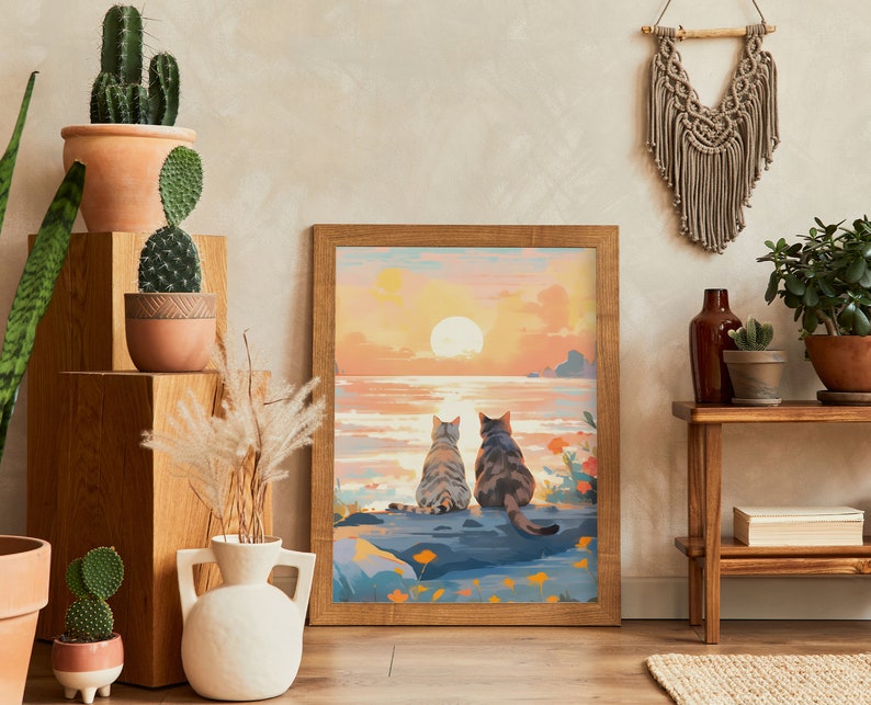 May include: A framed painting of two cats sitting on a rock by the ocean at sunset. The cats are looking out at the water. The painting is in a wooden frame and has a light brown background.