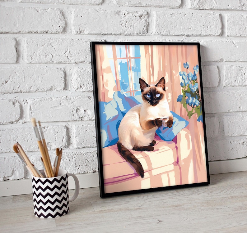 May include: Framed art print of a Siamese cat with blue eyes, sitting on a pink couch with blue pillows. The cat is holding a small cup. The background features a window and blue flowers.