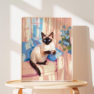 May include: A framed art print featuring a Siamese cat with blue eyes, holding a small cup. The cat is seated on a pink sofa with blue pillows, with a window and blue flowers in the background. The artwork has a soft, painterly style.