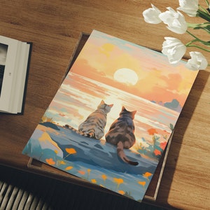 May include: Two gray tabby cats sit side-by-side on a rocky shore, looking out at a sunset over a body of water. The sky is a vibrant orange and pink, with a large sun setting in the distance.