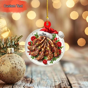 May include: A Christmas ornament featuring a plate of cooked lamb chops, decorated with tomatoes and greenery. The ornament is topped with a Santa hat and the name "Ann". The ornament is hanging from a gold string.
