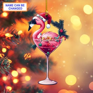 Könnte beinhalten: Rosafarbener Flamingo-Weihnachtsbaumschmuck mit roter Weihnachtsmannmütze, der ein Martini-Glas mit roter Flüssigkeit und Weihnachtsornamenten hält. Der Schmuck hat den Namen "Noah" auf der Mütze.