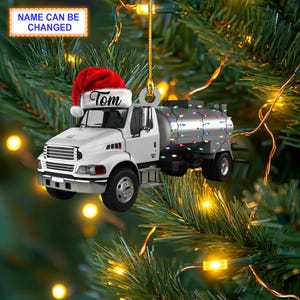 May include: A white Christmas ornament shaped like a tanker truck with a red Santa hat on top. The truck has a name "Tom" on the side. The truck is decorated with colorful Christmas lights.