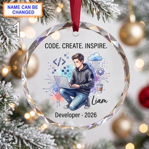 May include: A circular glass ornament with a red ribbon featuring an illustration of a person using a laptop, surrounded by digital icons, a cloud, and a coffee cup. Text reads: CODE. CREATE. INSPIRE. Liam Developer 2026.