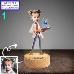 Puede incluir: Una luz nocturna personalizada con una ilustración de dibujos animados de una niña que lleva una bata de laboratorio y sostiene un libro. El nombre de la niña, "Lily", está escrito debajo de la ilustración. La base de la luz nocturna está hecha de madera y tiene las palabras "My Heart" grabadas.
