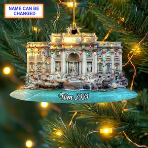 May include: A Christmas bauble featuring a colourful image of the Trevi Fountain in Rome, Italy. The fountain is decorated with twinkling lights and the text "Tom 2024" is printed below.