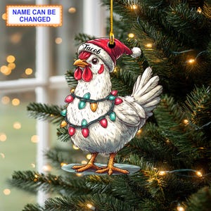 Puede incluir: Un pollo blanco con un gorro de Papá Noel rojo con el nombre "Jacob" en él. El pollo está decorado con luces de Navidad de colores y está de pie sobre una base blanca.