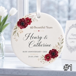 May include: A round white ceramic ornament hanging from a white ribbon, decorated with dark red floral illustrations and green leaves. The text reads 40 Beautiful Years, Henry & Catherine, RUBY ANNIVERSARY, 2026, and the date 24.05.1986.