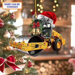 May include: Yellow and black toy construction vehicle ornament with a red Santa hat that says "Jack" on it. The ornament is decorated with a green wreath and gold bells. The vehicle is surrounded by a string of colorful Christmas lights.