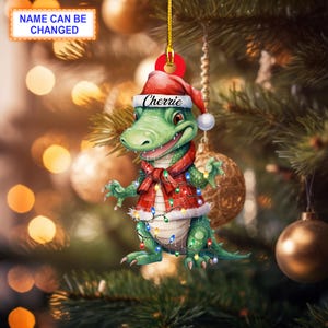 Op de afbeelding: Een groene alligator die een rode en witte kerstmanmuts en een rode geruite sjaal draagt met kerstverlichting eromheen gewikkeld. De ornament zegt "Cherrie" op de hoed.