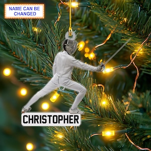 May include: A white Christmas ornament featuring a fencer in a white uniform holding a foil. The ornament is personalized with the name "CHRISTOPHER".