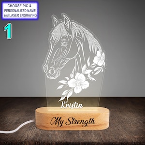 Pode incluir: Uma cabeça de cavalo branca com flores gravadas em um painel de acrílico transparente. O painel é montado em uma base de madeira com o nome "Kristin" e a frase "My Strength" gravadas nela. A base tem um cabo branco conectado a ela.