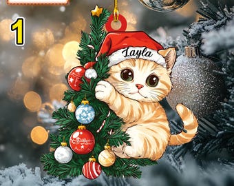 May include: A Christmas ornament featuring a cartoon cat wearing a Santa hat and holding onto a small Christmas tree decorated with ornaments. The cat's name, "Layla", is written on the hat.