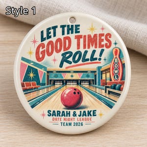 May include: A round ceramic ornament featuring a bowling alley illustration with a red bowling ball in the foreground. The text reads LET THE GOOD TIMES ROLL! SARAH & JAKE DATE NIGHT LEAGUE TEAM 2026. It includes a small hole at the top for hanging.