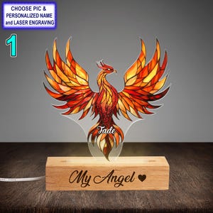 Puede incluir: Un fénix de vitral con colores naranja, amarillo y rojo se ilumina desde atrás. La base de la lámpara es de madera y tiene las palabras "My Angel" grabadas.