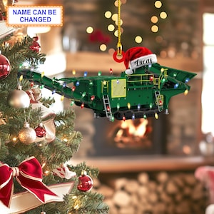 Puede incluir: Un adorno navideño verde con forma de trituradora de roca con un gorro de Papá Noel rojo en la parte superior. El adorno tiene una cadena de luces de colores envuelta alrededor y el nombre "Lucas" está escrito en el gorro.