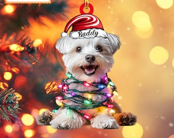 May include: A white dog with a red Santa hat that says "Buddy" on it is wrapped in colorful Christmas lights. The dog is sitting on a blurry background of a Christmas tree with lights.