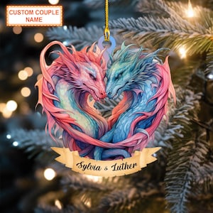 May include: A heart-shaped Christmas ornament featuring two intertwined dragons, one pink and one blue. The dragons are facing each other with their heads touching. The ornament has a gold ribbon for hanging and a banner with the text "Sylvia & Luther".