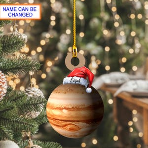 May include: A Christmas ornament shaped like the planet Jupiter, with a red Santa hat and the name "Elijah" on it. The ornament hangs from a gold string. The text "NAME CAN BE CHANGED" is in a blue box.