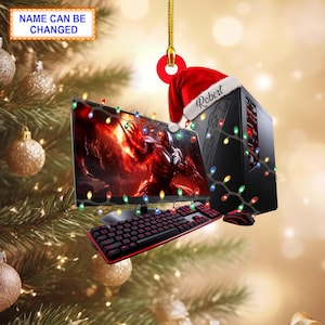 Puede incluir: Un adorno navideño con forma de torre de ordenador negra con un gorro de Papá Noel rojo que dice "Robert". La torre está decorada con una cadena de luces navideñas de colores. También se incluye un teclado y un monitor de ordenador negros en el adorno.