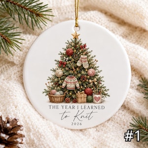Könnte beinhalten: Ein rundes, weißes Keramikornament mit einem Weihnachtsbaum, der mit Wollknäueln, einem Miniaturpullover, Fäustlingen und Stricknadeln geschmückt ist. Unter dem Baum steht der Text THE YEAR I LEARNED to Knit 2026. Es hängt an einer goldenen Schnur.