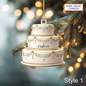 Könnte beinhalten: Ein dreistufiger weißer Kuchenschmuck mit goldenen Verzierungen und perlenartigen Akzenten. Der Schmuck hat das Wort "Custom" auf der mittleren Ebene und den Text "NAME CAN BE CHANGED" auf einem kleinen Schild. Der Schmuck ist mit "Style 1" gekennzeichnet.