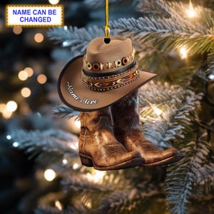 Puede incluir: Un sombrero de vaquero marrón con una banda de cuentas y un par de botas de vaquero marrones. El sombrero tiene el texto "Name Here" bordado. El adorno está colgado de un árbol de Navidad.
