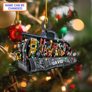 Peut inclure: Un sac à outils noir avec le nom personnalisé "DAVID" sur le devant. Le sac est rempli d'outils et décoré de lumières de Noël colorées.