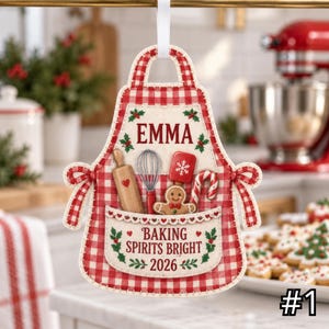 Op de afbeelding: Een rood-wit geblokt schortvormig ornament met de naam EMMA, een deegroller, garde, spatel, peperkoekmannetje en zuurstok. De tekst luidt BAKING SPIRITS BRIGHT 2026, versierd met hulstblaadjes en bessen op een witte achtergrond.