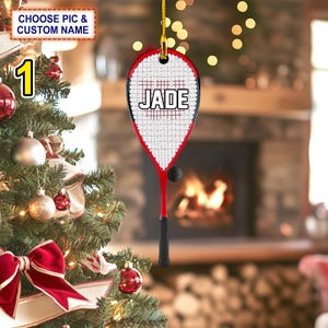 May include: Red and black squash racket Christmas ornament with a white ball and the name "JADE" printed on the racket.