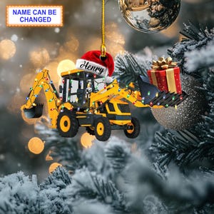 May include: Yellow toy backhoe with Christmas lights, a red Santa hat with the name "Henry" on it, and a red gift box. The backhoe is hanging from a gold string, and the background is a snowy Christmas tree.