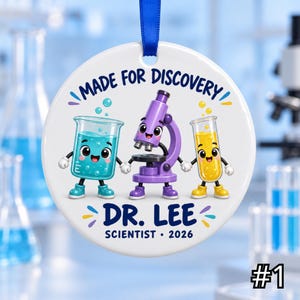 Op de afbeelding: Een rond wit keramieken ornament met een blauw lint. Het ontwerp toont een cartoon bekerglas, microscoop en reageerbuis met gezichtjes die elkaars hand vasthouden. Tekst: MADE FOR DISCOVERY, DR. LEE, SCIENTIST, 2026. Kleuren: blauw, paars en geel.