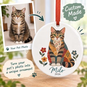 Peut inclure: Une décoration ronde en céramique blanche suspendue par un ruban rouge, illustrée d'un chat tigré au style patchwork assis parmi des fleurs. Le prénom Milo et l'année 2026 sont inscrits sous le chat dans une police cursive bleu sarcelle.