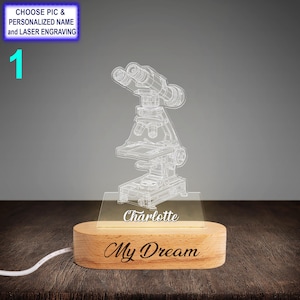 Puede incluir: Un diseño de microscopio blanco impreso en 3D sobre una base de acrílico transparente con una base de madera. La base tiene el nombre "Charlotte" y el texto "My Dream" grabado.