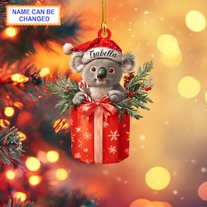 May include: A Christmas ornament featuring a gray koala wearing a red Santa hat, peeking out of a red and white gift box with a red bow. The ornament has the name "Isabella" written on the hat.